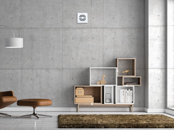 Modern eingerichtetes Wohnzimmer mit einem braunen Sessel, Regal und einer Steuerungseinheit an der Betonwand | © bösch heizung.klima.lüftung