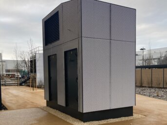 Modernes, schwarz-weiß gestreiftes Lüftungsgerät zur Vorfilterung von Außenluft auf einem Kiesbett in einem urbanen Umfeld. Das Gerät steht frei auf einer gepflasterten Fläche, umgeben von einer niedrigen Mauer und einem Zaun, mit industriellen Strukturen im Hintergrund. | © Walter Bösch GmbH & Co KG