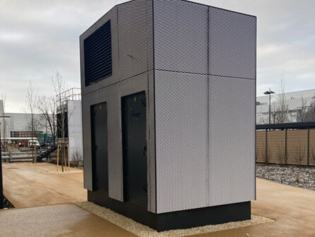 Modernes, schwarz-weiß gestreiftes Lüftungsgerät zur Vorfilterung von Außenluft auf einem Kiesbett in einem urbanen Umfeld. Das Gerät steht frei auf einer gepflasterten Fläche, umgeben von einer niedrigen Mauer und einem Zaun, mit industriellen Strukturen im Hintergrund. | © Walter Bösch GmbH & Co KG