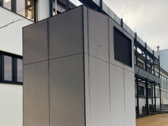 Hohes, modernes Lüftungsgerät zur Vorfilterung von Außenluft, ummantelt mit schwarz-weißem Gitter, steht auf einem gepflasterten Boden vor einem mehrstöckigen Gebäude mit großen Fenstern. Das Gerät ist an der Basis mit einem schwarzen Rand versehen und von einer Schicht Kies umgeben. | © Walter Bösch GmbH & Co KG