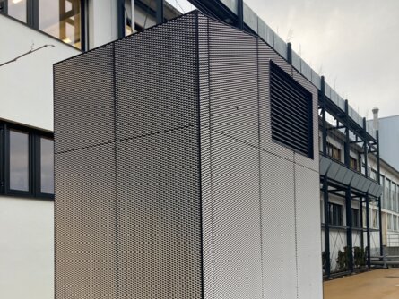 Hohes, modernes Lüftungsgerät zur Vorfilterung von Außenluft, ummantelt mit schwarz-weißem Gitter, steht auf einem gepflasterten Boden vor einem mehrstöckigen Gebäude mit großen Fenstern. Das Gerät ist an der Basis mit einem schwarzen Rand versehen und von einer Schicht Kies umgeben. | © Walter Bösch GmbH & Co KG