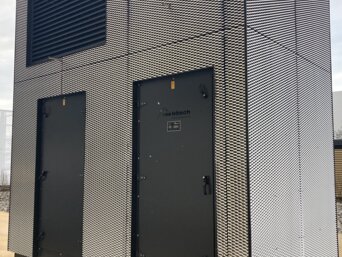 Vertikales Lüftungsgerät zur Vorfilterung von Außenluft, ummantelt mit einem feinmaschigen schwarz-weißen Gitter, steht auf einem Kiesbett. Das Gerät besitzt zwei schwarze Türen und ein großes Lüftungsgitter oben. Ein Firmenlogo ist sichtbar an der Front des Geräts. | © Walter Bösch GmbH & Co KG