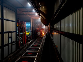 Innenansicht einer Entstaubungsanlage. Im Vordergrund links sieht man die Außenwand der Einhausung mit Gitterstrukturen und eine offene Tür. Rechts im Bild ist ein langer Tunnel sichtbar, in dem die Gießstraße installiert ist. Am Ende des Tunnels sind Lichter und Arbeiter zu erkennen. Der Fokus liegt auf einem beleuchteten Bereich in der Mitte des Tunnels. | © INFRANORM®