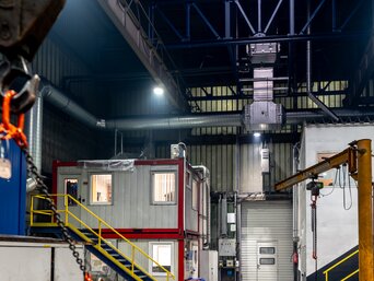 Innenansicht einer Industriehalle mit einer nachhaltigen Hallenklimatisierung. Im Vordergrund links hängt eine orange Kette mit einem Haken, die von einem Kran herabhängt. Rechts im Bild ist ein modularer Bürocontainer in Rot und Grau auf zwei Ebenen zu sehen, der auf einer gelben Stahlkonstruktion steht. Darüber befindet sich eine moderne Lüftungs- und Beleuchtungsinstallation an der Hallendecke. | © INFRANORM®