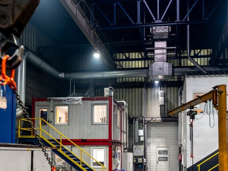 Innenansicht einer Industriehalle mit einer nachhaltigen Hallenklimatisierung. Im Vordergrund links hängt eine orange Kette mit einem Haken, die von einem Kran herabhängt. Rechts im Bild ist ein modularer Bürocontainer in Rot und Grau auf zwei Ebenen zu sehen, der auf einer gelben Stahlkonstruktion steht. Darüber befindet sich eine moderne Lüftungs- und Beleuchtungsinstallation an der Hallendecke. | © INFRANORM®