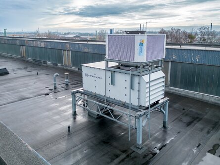 Luftaufnahme eines großen, auf einem Fabrikdach installierten Lüftungssystems. Die Anlage steht auf einer Metallstruktur und ist vorwiegend in Weiß gehalten, mit einem oberen Gitter in Silber. Das Dach ist nass, möglicherweise nach einem Regenschauer. Im Hintergrund ist eine städtische Umgebung mit entfernten Gebäuden und leicht bewölktem Himmel zu erkennen. | © INFRANORM®