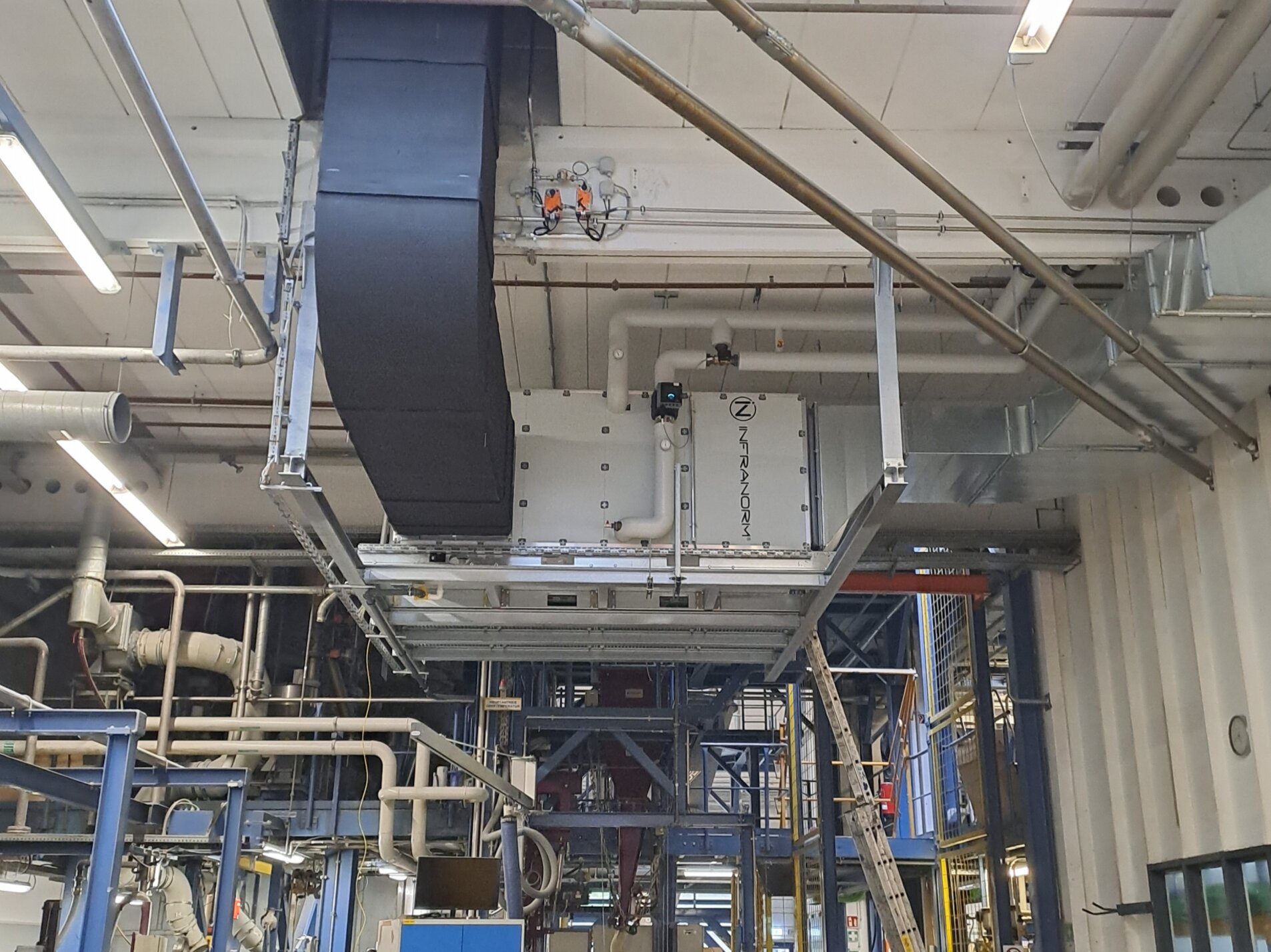 Industrielle Produktionshalle mit komplexen Rohrleitungen und einer großen, schwarzen Lüftungsanlage an der Decke. Unten arbeitet eine Person an einem Tisch mit einem Laptop, umgeben von Maschinen und technischen Geräten. Im Hintergrund sind Leitern und weitere technische Strukturen zu sehen. | © Infranorm, Wels (AT)