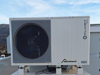 Große, industrielle Klimaanlage der Marke Clivet mit dem Logo von bösch auf der Vorderseite, auf einem Dach installiert. Die Anlage ist grau und hat einen großen, kreisförmigen Lüfter auf der linken Seite. Im Hintergrund sind Berge und ein leicht bewölkter Himmel sichtbar.