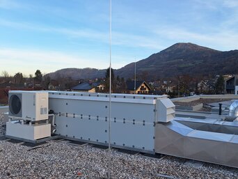 Weitwinkelaufnahme eines Daches bedeckt mit Kies und mehreren Klimaanlagen-Einheiten. Das Dach bietet eine Aussicht auf eine hügelige Landschaft mit Bäumen und einige Gebäude im Hintergrund unter einem klaren Abendhimmel.