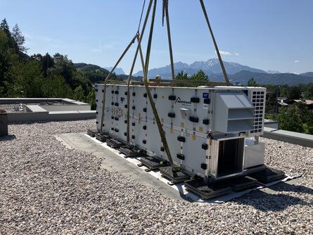 Großes, weißes modulares Lüftungsgerät wird auf einem kiesbedeckten Dach installiert, umgeben von Seilen und Hebezeug. Im Hintergrund sind bewaldete Berge unter einem sonnigen blauen Himmel sichtbar.