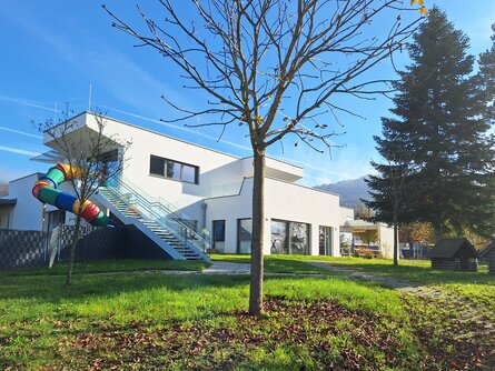 Modernes weißes Kindergarten-Gebäude mit bunter Rutsche, die an der Außenseite angebracht ist. Im Vordergrund steht ein einzelner Baum auf einem Rasen mit herbstlich gefärbten Blättern auf dem Boden, unter einem klaren blauen Himmel. | © Marius Projekt ZT GmbH