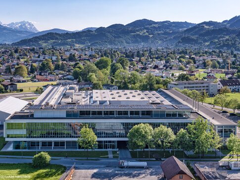 Luftaufnahme eines modernen Gebäudekomplexes mit Pv-Anlagen auf dem Dach, umgeben von einer idyllischen Landschaft und Bergen im Hintergrund | © Lukas Hämmerle