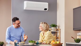 Ein Paar sitzt in einer modernen Küche, entspannt bei einer Mahlzeit, während ein Samsung WindFree Klimagerät an der Wand für ein angenehmes Raumklima sorgt. | © Samsung