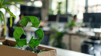 Grünes Recycling-Symbol in einem Karton vor unscharfem Bürohintergrund.